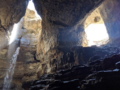 A cavern with two openings at the top, sunlight streaming in, and a waterfall cascading down rocky walls.