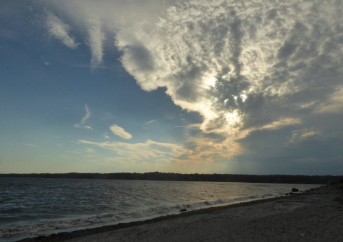 A serene beach scene with a cloudy sky and sunlight peeking through, reflecting on the calm water.