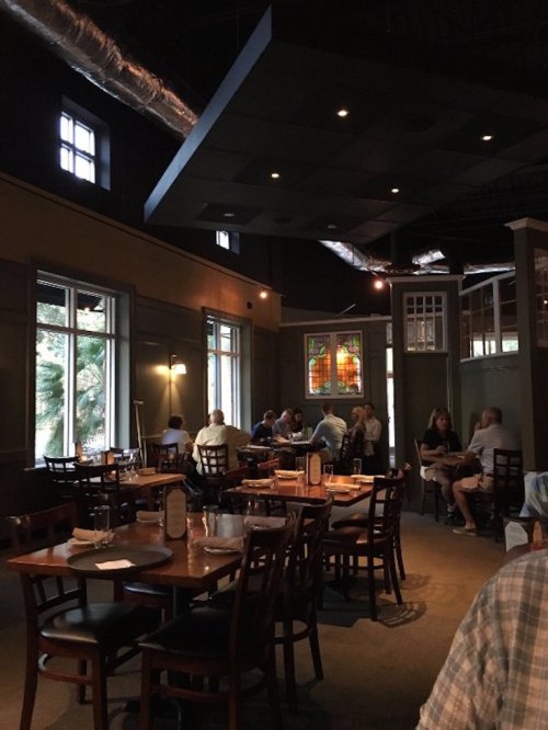 Cozy restaurant interior with wooden tables, chairs, and soft lighting; patrons dining and socializing.