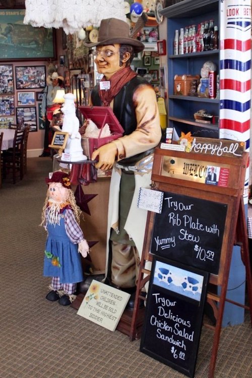 A cowboy statue stands beside a menu board and a scarecrow in a cozy restaurant filled with vintage decor.