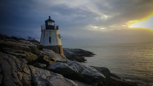 A lighthouse stands on rocky shores, overlooking a calm sea under a cloudy sky at sunset.