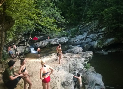 A group of people enjoying a sunny day by a rocky riverbank, some swimming and others relaxing in nature.