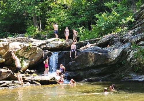 A group of people enjoying a sunny day by a rocky stream, swimming and relaxing near a small waterfall.
