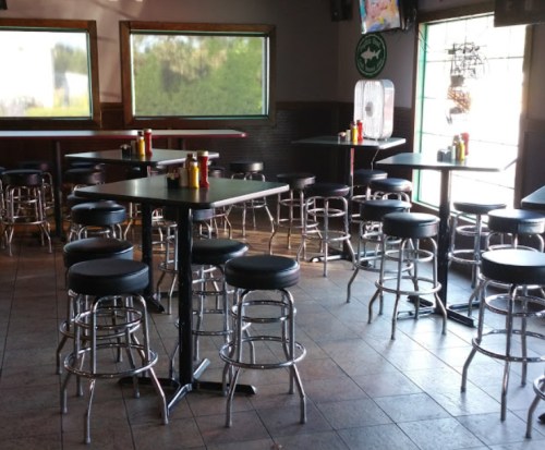 Empty bar area with black stools and tables, featuring condiments on each table and large windows letting in light.