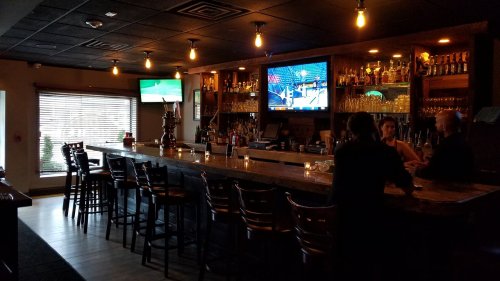 A dimly lit bar with a long counter, shelves of liquor, and two TVs showing sports, with a few patrons seated.