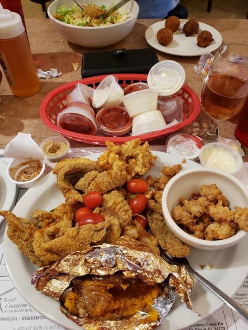 A table filled with fried food, including fish, tomatoes, and sides, along with drinks and a salad in the background.