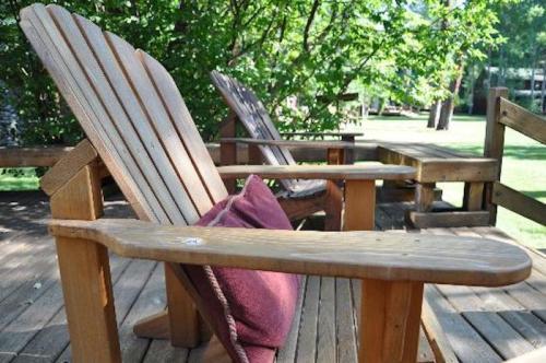 Two wooden Adirondack chairs on a deck surrounded by greenery, with a cushion on one chair.