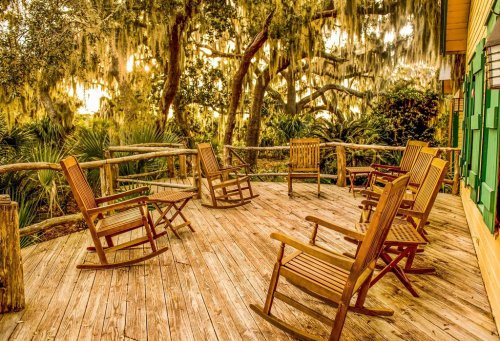 A wooden deck with rocking chairs surrounded by lush greenery and trees draped in Spanish moss, bathed in warm light.