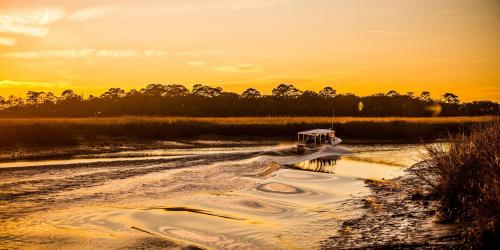 A boat glides through a calm river at sunset, surrounded by tall grasses and trees under a golden sky.