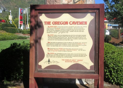 Sign for "The Oregon Cavemen," detailing the group's history and activities, surrounded by greenery and a scenic backdrop.