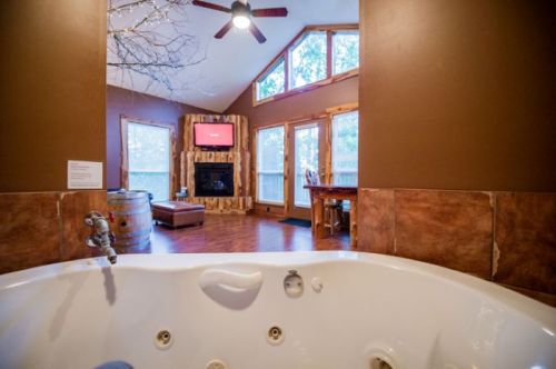 A cozy interior featuring a hot tub, wooden accents, large windows, and a view of a living area with a fireplace.