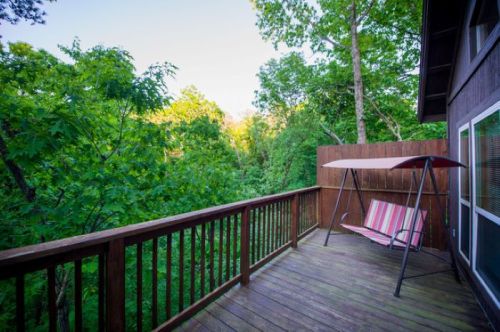 A wooden deck with a swing chair, surrounded by lush green trees and a clear sky in the background.