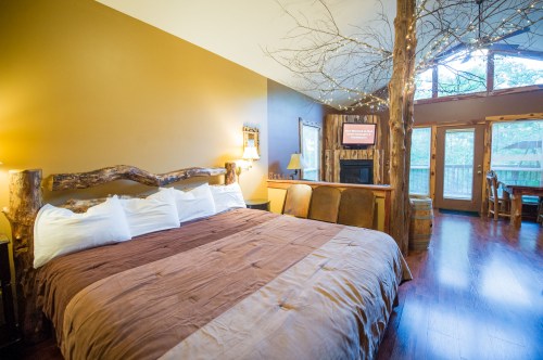 Cozy bedroom with a large bed, wooden accents, and a view of nature through glass doors. Warm lighting creates a welcoming atmosphere.