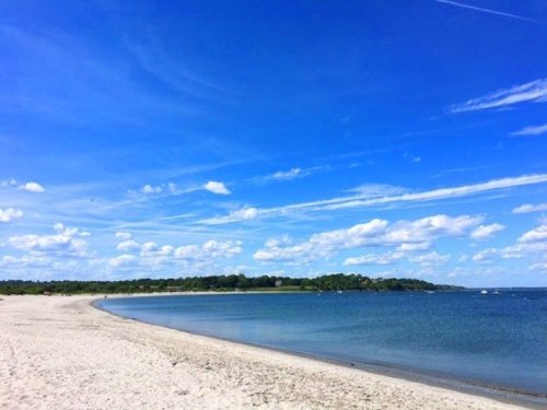 A serene beach scene with soft sand, calm water, and a clear blue sky dotted with fluffy clouds.
