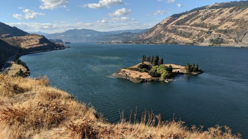 A scenic view of a river winding through hills, with an island featuring trees in the foreground and mountains in the background.