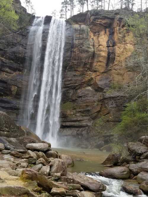 A stunning waterfall cascades down rocky cliffs, surrounded by lush greenery and mist rising from the water below.