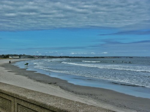 A serene beach scene with gentle waves, a cloudy sky, and a surfer approaching the water along a sandy shore.