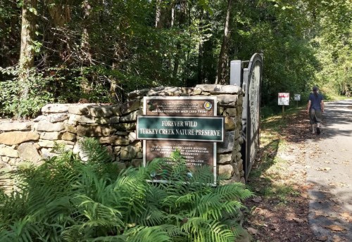 A stone entrance sign for Turkey Creek Nature Preserve, surrounded by trees and ferns, with a person walking nearby.