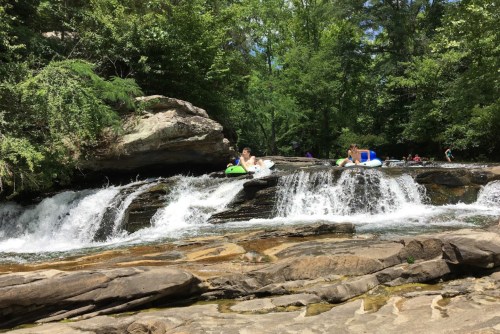People enjoying a sunny day by a waterfall, with some sitting on rocks and others in inflatable tubes.