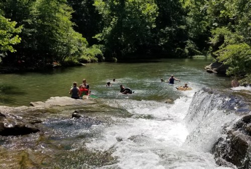 People swimming and relaxing in a natural river surrounded by lush greenery and a small waterfall.
