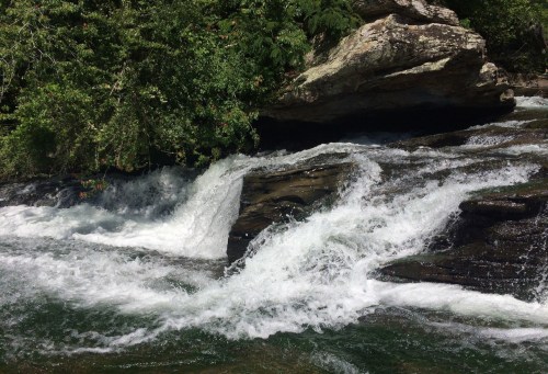 A rushing river cascades over rocks, surrounded by lush greenery and sunlight filtering through the trees.