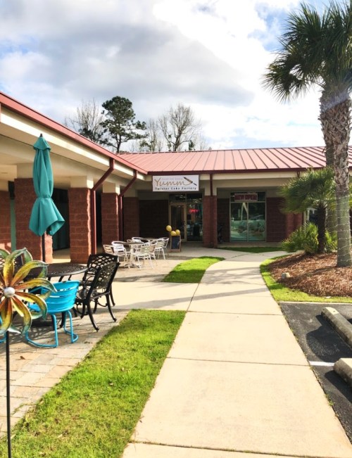 A pathway leads to a restaurant entrance, with outdoor seating and palm trees nearby under a partly cloudy sky.