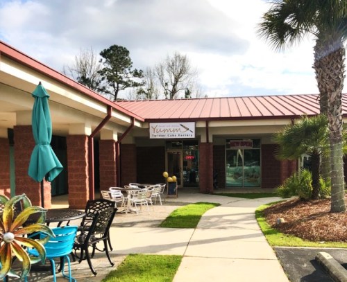 Exterior view of a cafe with a sign reading "Yumm," featuring outdoor seating and palm trees.