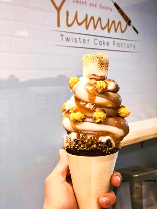 A hand holding a tall ice cream cone topped with caramel, chocolate, popcorn, and a marshmallow.