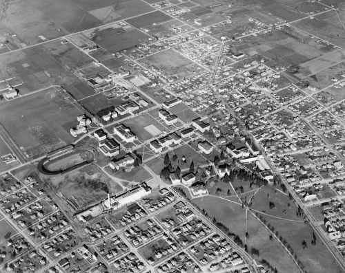 Aerial view of a town with a mix of residential areas, fields, and institutional buildings, in black and white.