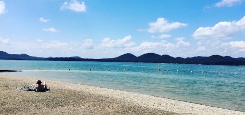 A person relaxes on a beach by a calm lake, surrounded by mountains and a blue sky with fluffy clouds.