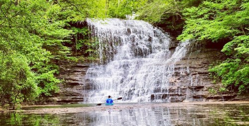 A serene waterfall cascades over rocks, surrounded by lush greenery, with a person sitting peacefully in the foreground.