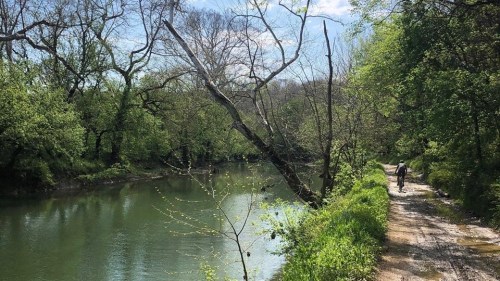 A peaceful river scene with lush greenery and a person walking along a dirt path beside the water.