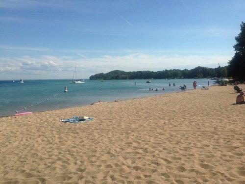 A sandy beach with people swimming and relaxing, clear blue water, and a few boats in the distance under a sunny sky.