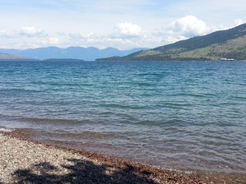 A serene lake with gentle waves, surrounded by mountains and a clear blue sky, viewed from a rocky shoreline.