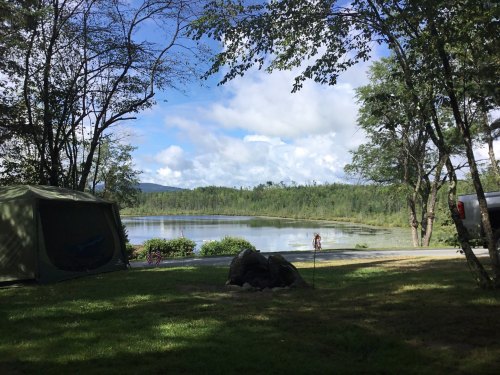A serene lakeside view with a tent, trees, and a person walking along the water under a partly cloudy sky.