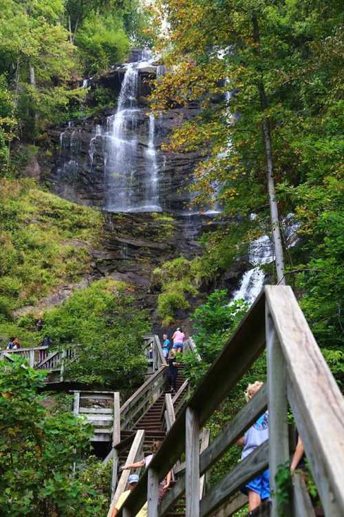 A scenic waterfall cascades down rocky cliffs, surrounded by lush greenery and wooden steps leading up the hillside.