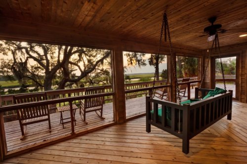 A cozy wooden porch with a swing, rocking chairs, and a view of trees and a serene landscape at sunset.