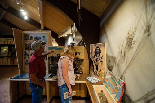 Two visitors explore an exhibit in a museum, examining historical photographs and artifacts displayed on wooden panels.