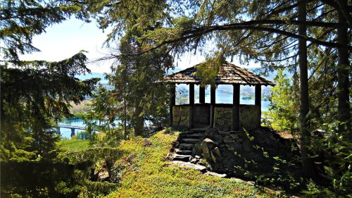 A small gazebo nestled among trees, overlooking a serene body of water and distant landscape under a clear blue sky.