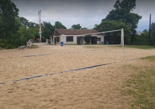 A sandy volleyball court with nets, next to a small building surrounded by trees under a cloudy sky.