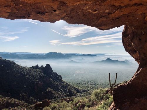 View from a rocky cave opening overlooking a misty valley and distant mountains under a clear blue sky.