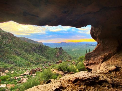 A scenic view of a mountainous landscape framed by a rocky cave opening, with vibrant skies at sunset.