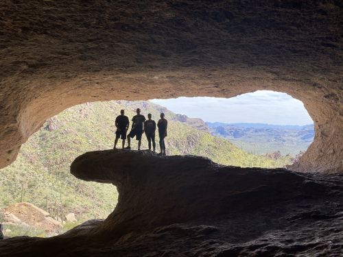 Four silhouetted figures stand on a rock ledge inside a cave, framed by a large opening with a scenic landscape beyond.
