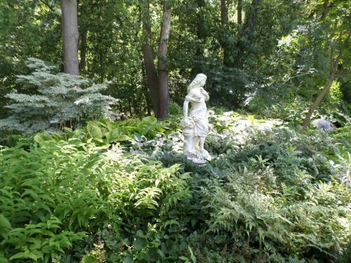 A white statue of a woman stands amidst lush green foliage in a serene garden setting.