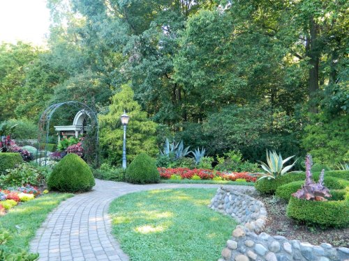 A winding stone path through a lush garden with colorful flowers, shrubs, and a decorative archway in the background.