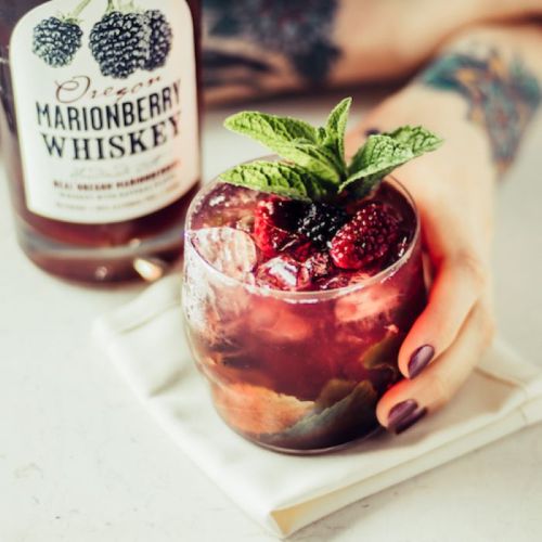 A hand holds a glass of marionberry whiskey cocktail, garnished with mint and berries, next to a bottle of whiskey.