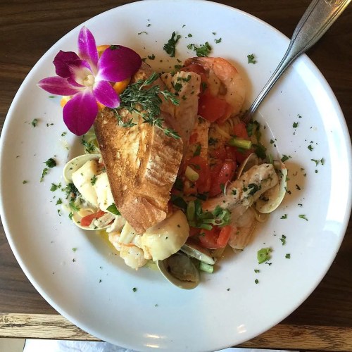 A plate of seafood pasta with shrimp, clams, and a slice of toasted bread, garnished with herbs and an orchid.