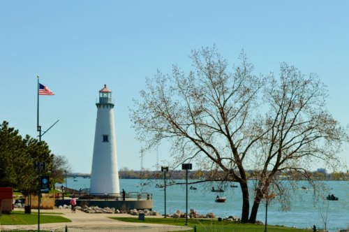 A white lighthouse beside a lake, with a tree and boats in the water, under a clear blue sky. An American flag is visible.