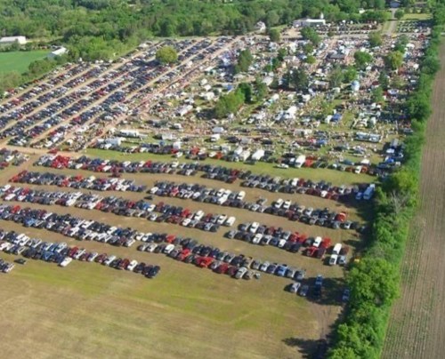 Aerial view of a large outdoor event with rows of parked cars and green fields surrounding the area.
