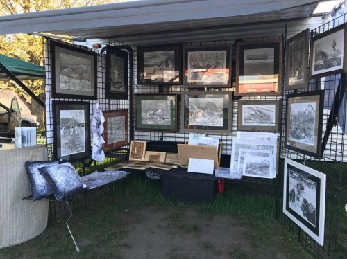 Art display featuring various framed drawings and prints, arranged on a grid wall under a canopy.